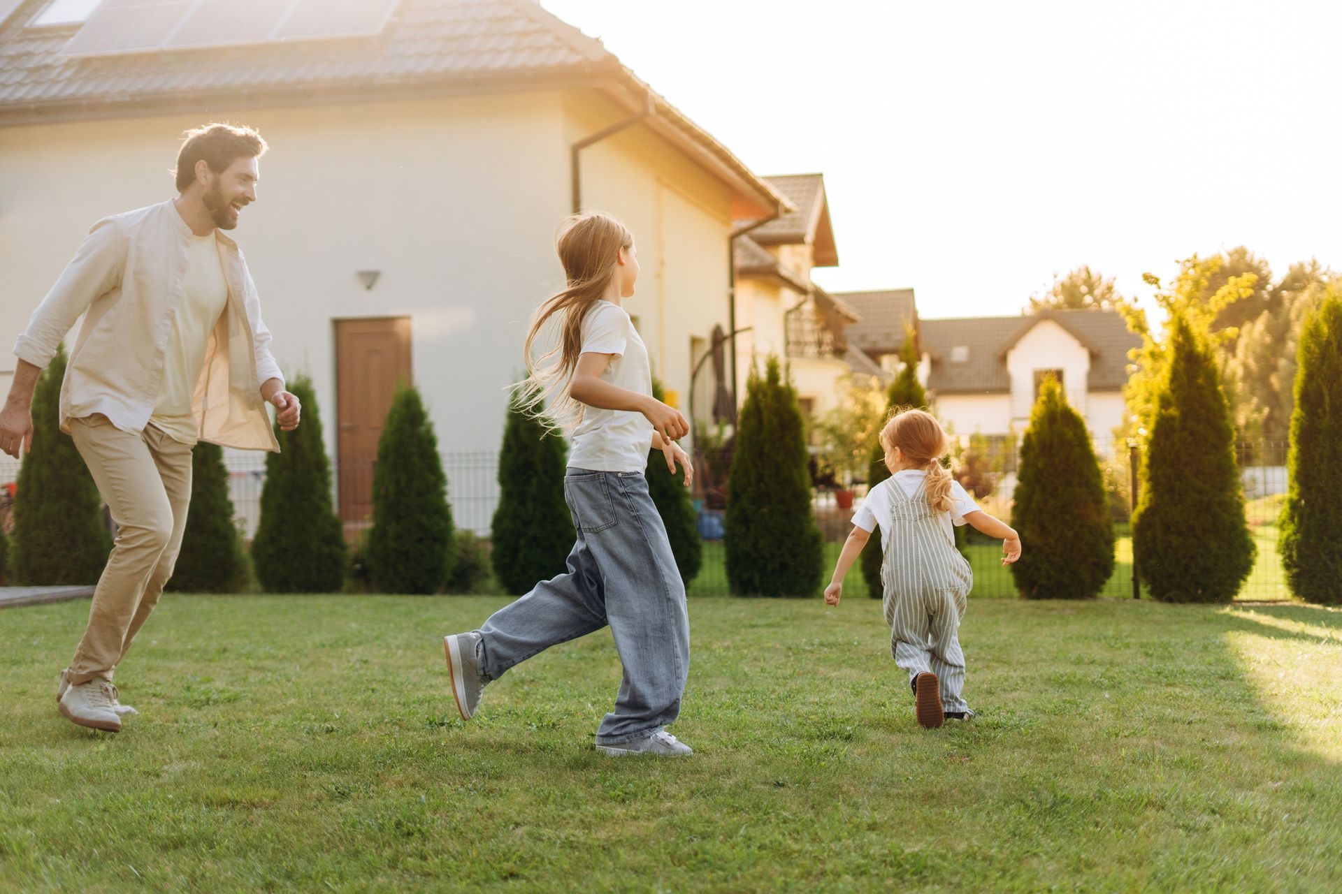 A person playing and running on a green lawn with two children in front of a house at sunset.