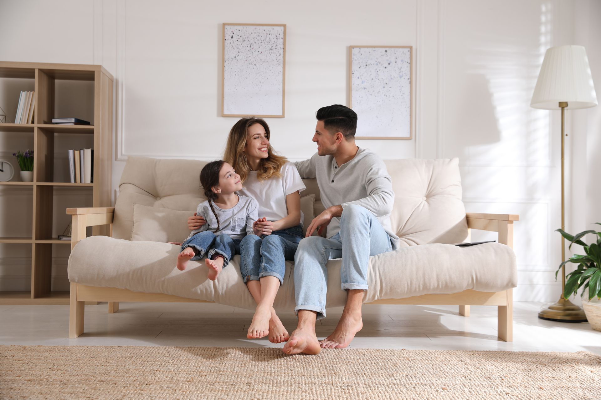 A family sits together on a light-colored sofa in a bright living room, smiling and talking to each other.