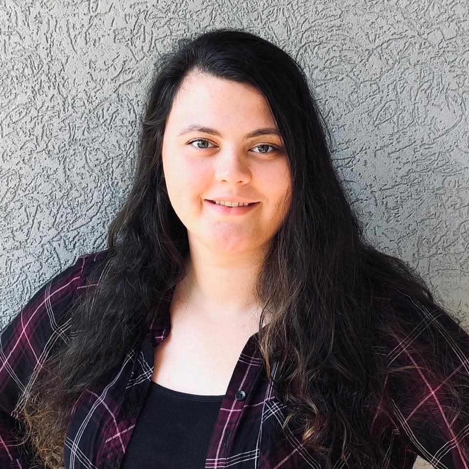 A person with long dark hair, wearing a black and purple plaid shirt over a black top, posing against a textured grey wall.