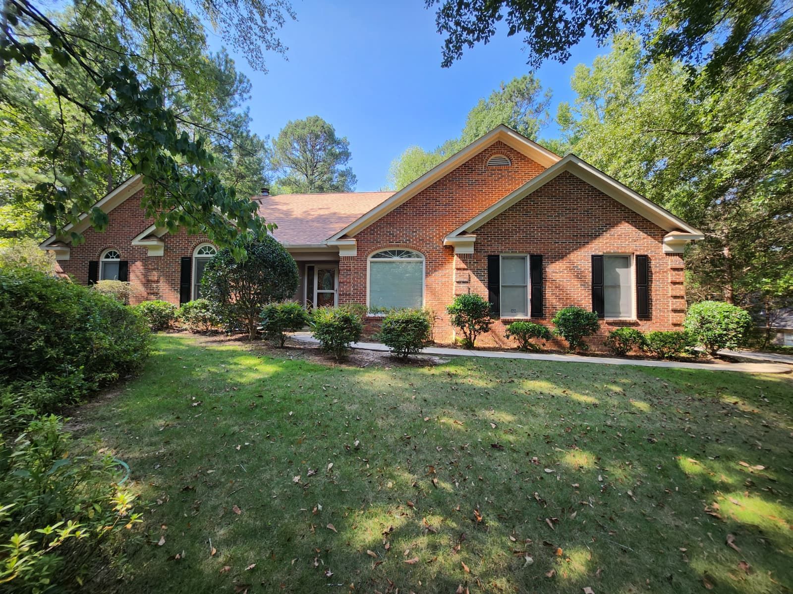 Single-story brick house with a front lawn, surrounded by mature green trees on a sunny day.