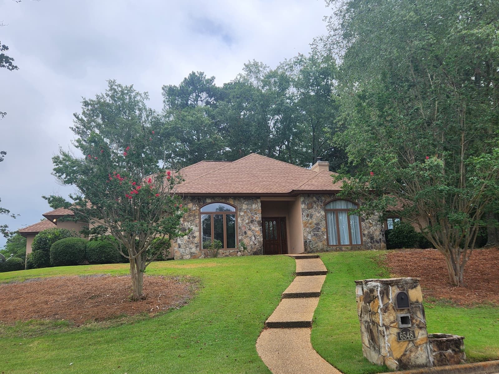 A stone house with a red tile roof and a pebble walkway leading to the front door, surrounded by trees and green lawn.