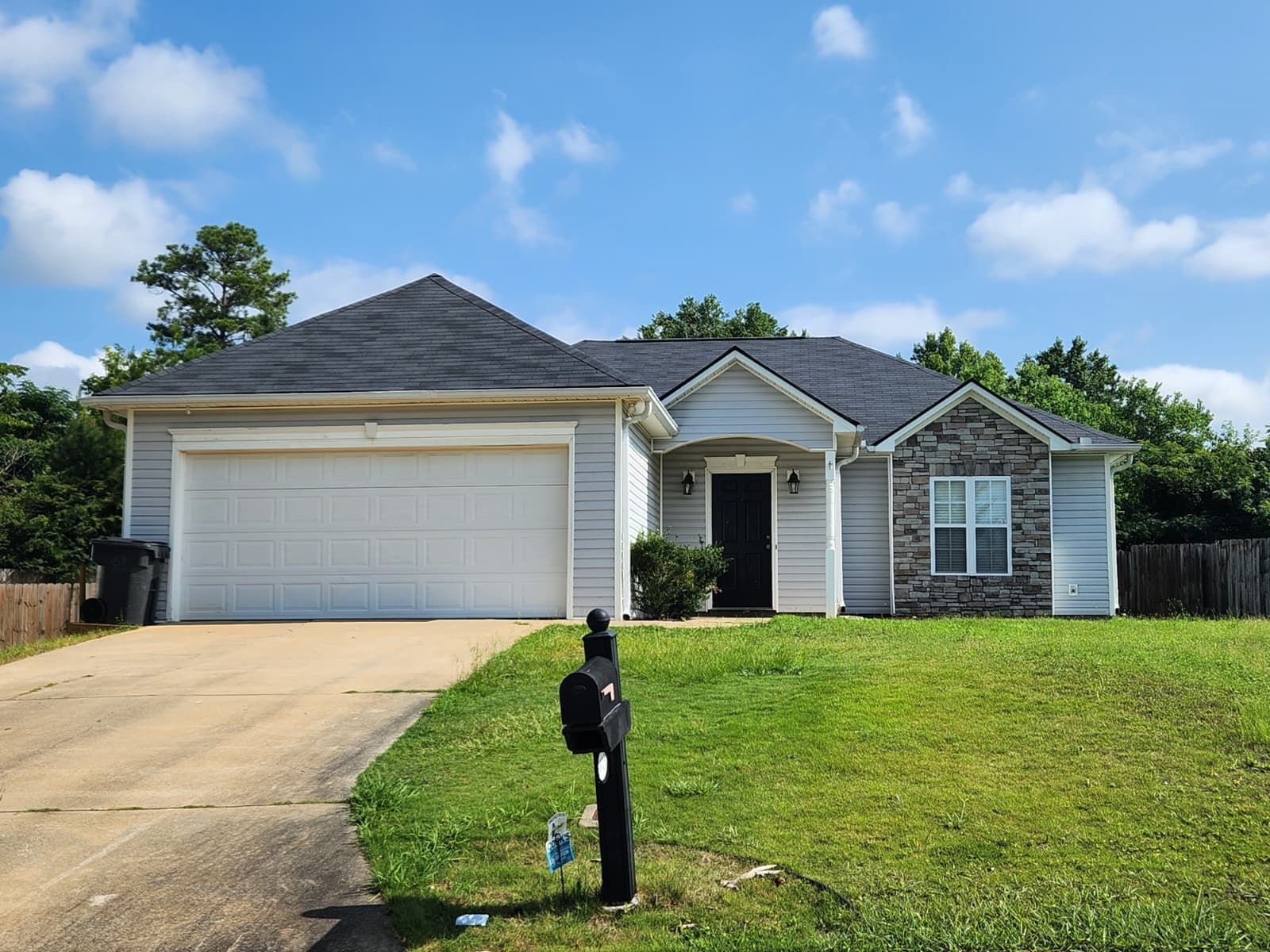 A single-story suburban house with a two-car garage, light-colored siding, stone accents, and a dark roof under a blue sky.