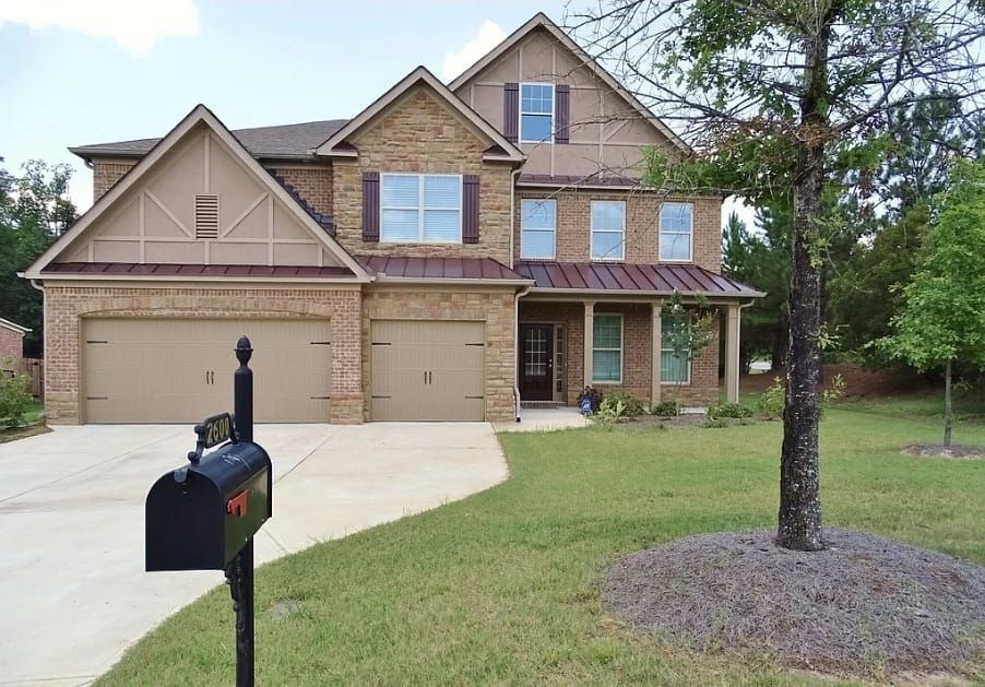 A two-story tan brick suburban house with a two-car garage and a metal roof, fronted by a lawn and a black mailbox.