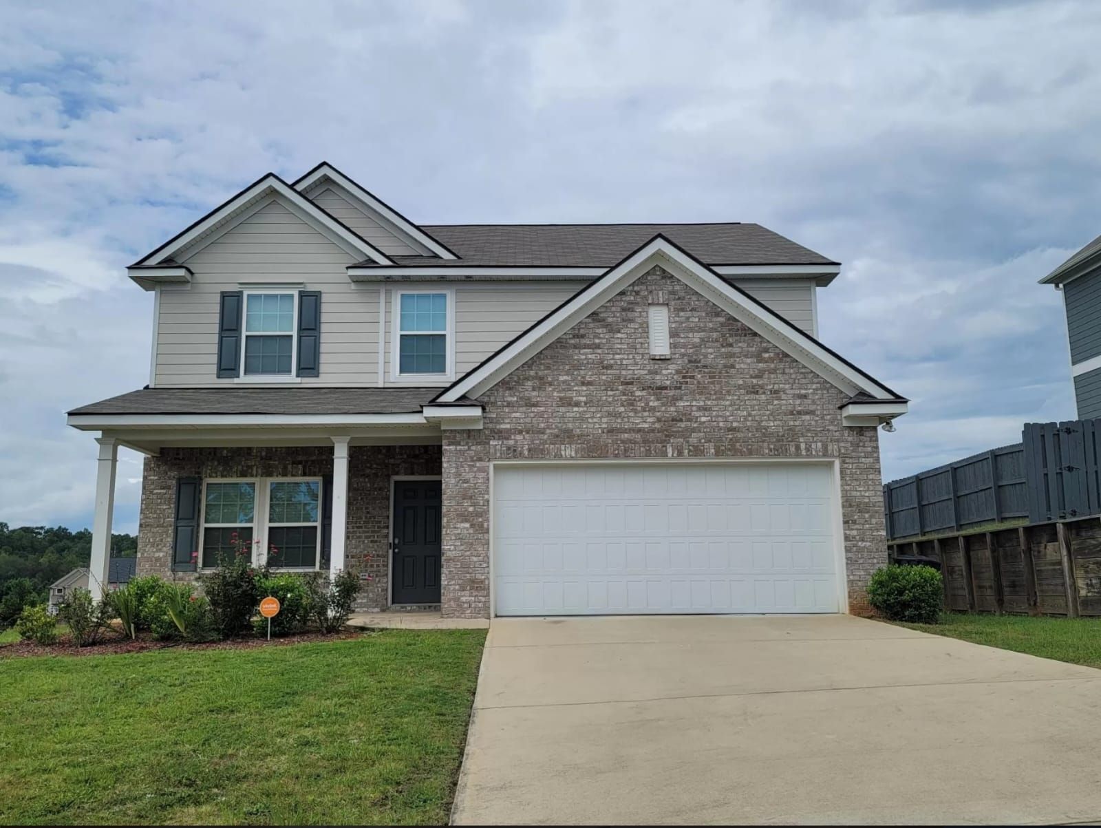 A two-story suburban home with a gray brick facade, light siding, a white garage door, and a front lawn under a cloudy sky.