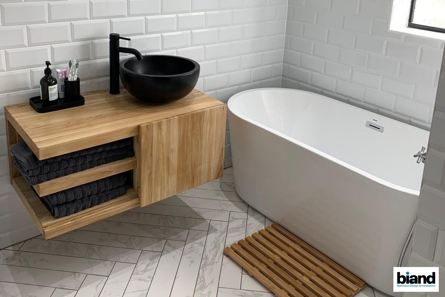 Modern bathroom with wooden vanity, black sink, white tub, and tiled walls.