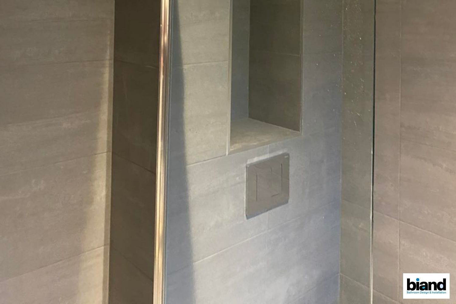 Gray tiled shower with built-in shelf and flush-mount fixture; glass door on the left.