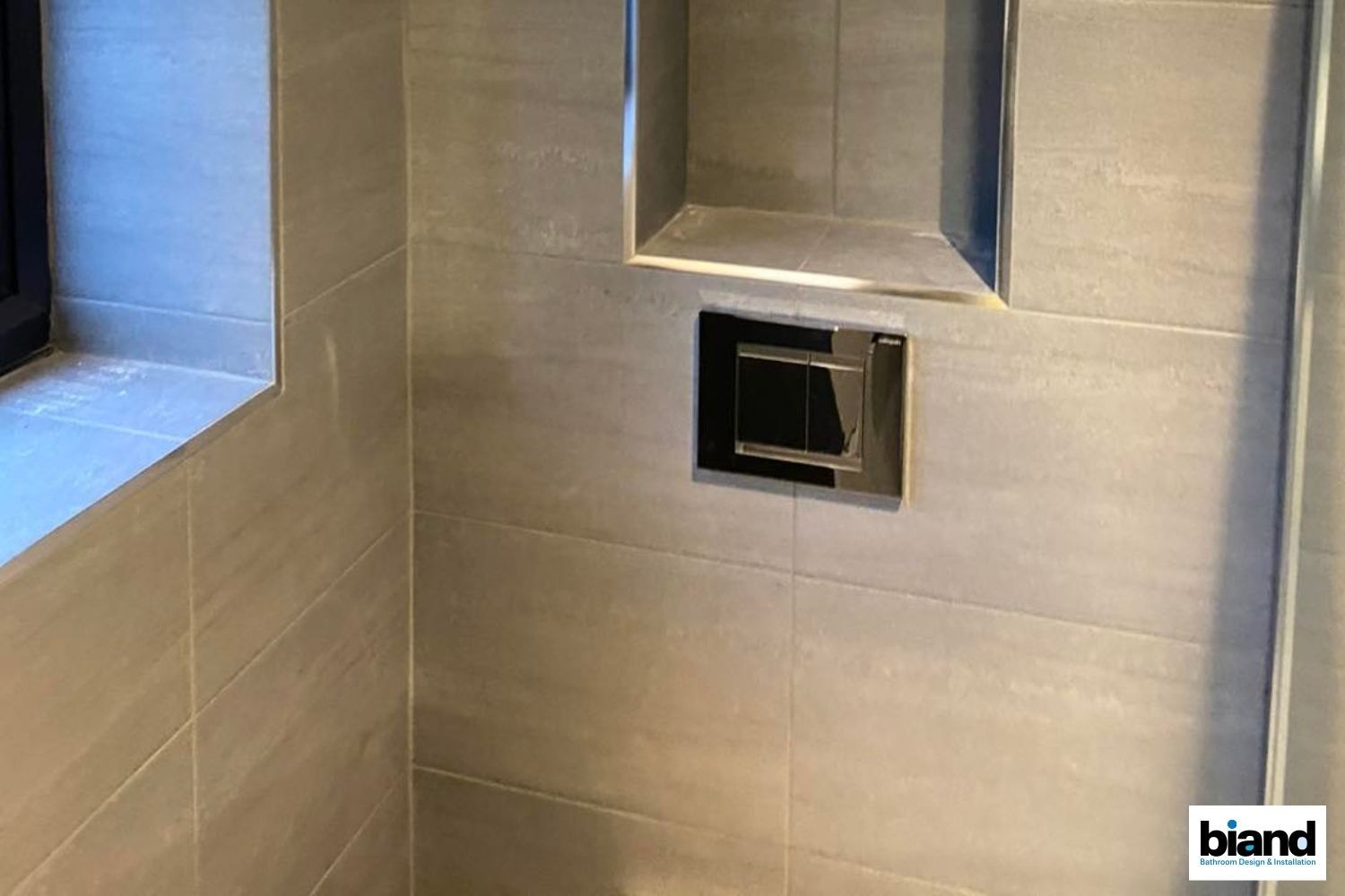 Bathroom wall with gray tiles, built-in shelf, and a chrome flush plate.