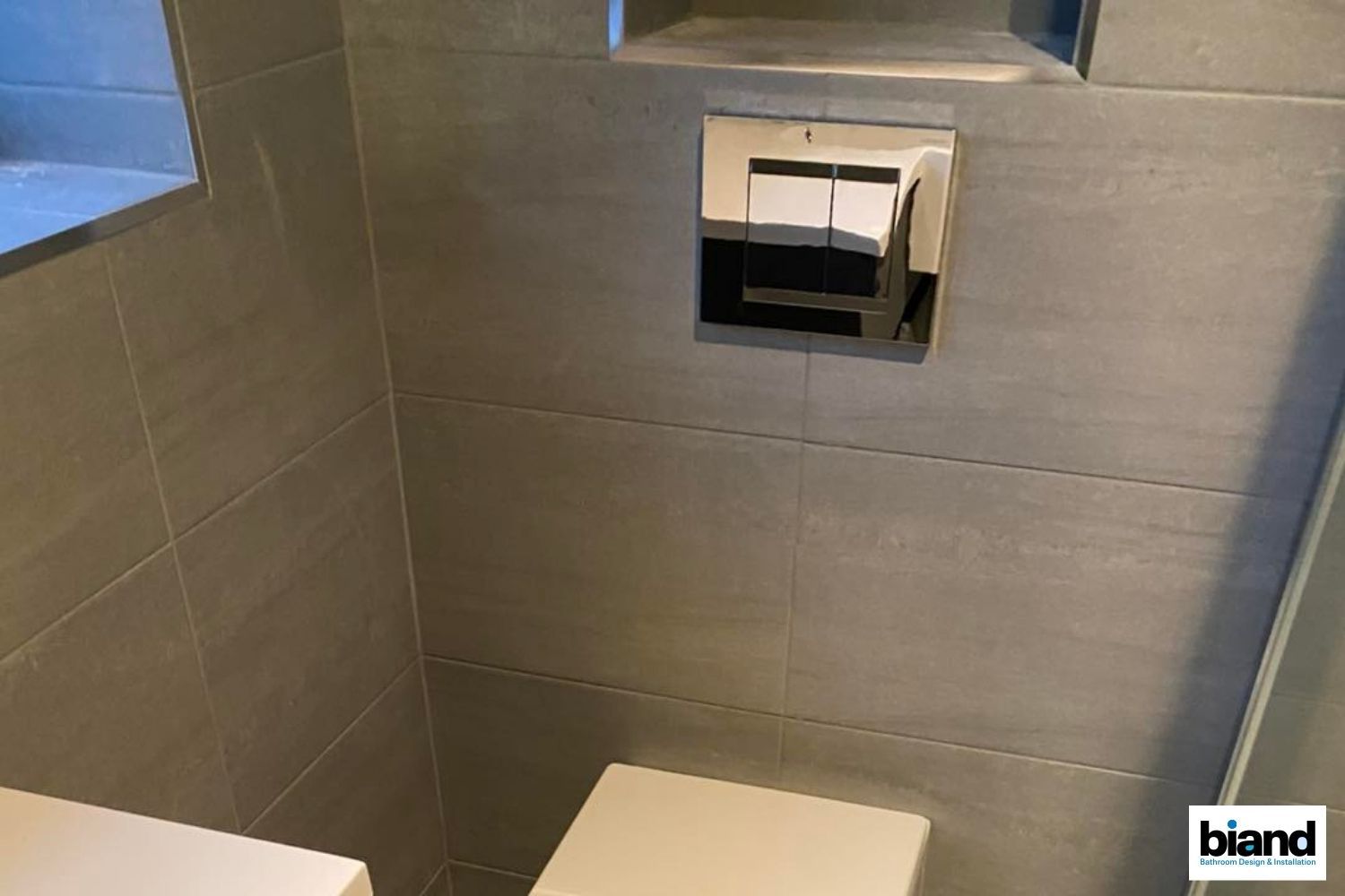 A modern, gray-tiled bathroom with a chrome flush plate, and a white toilet seat.