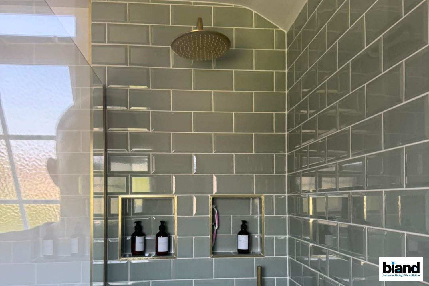 Gray tiled shower with gold showerhead, recessed shelves holding dark bottles.