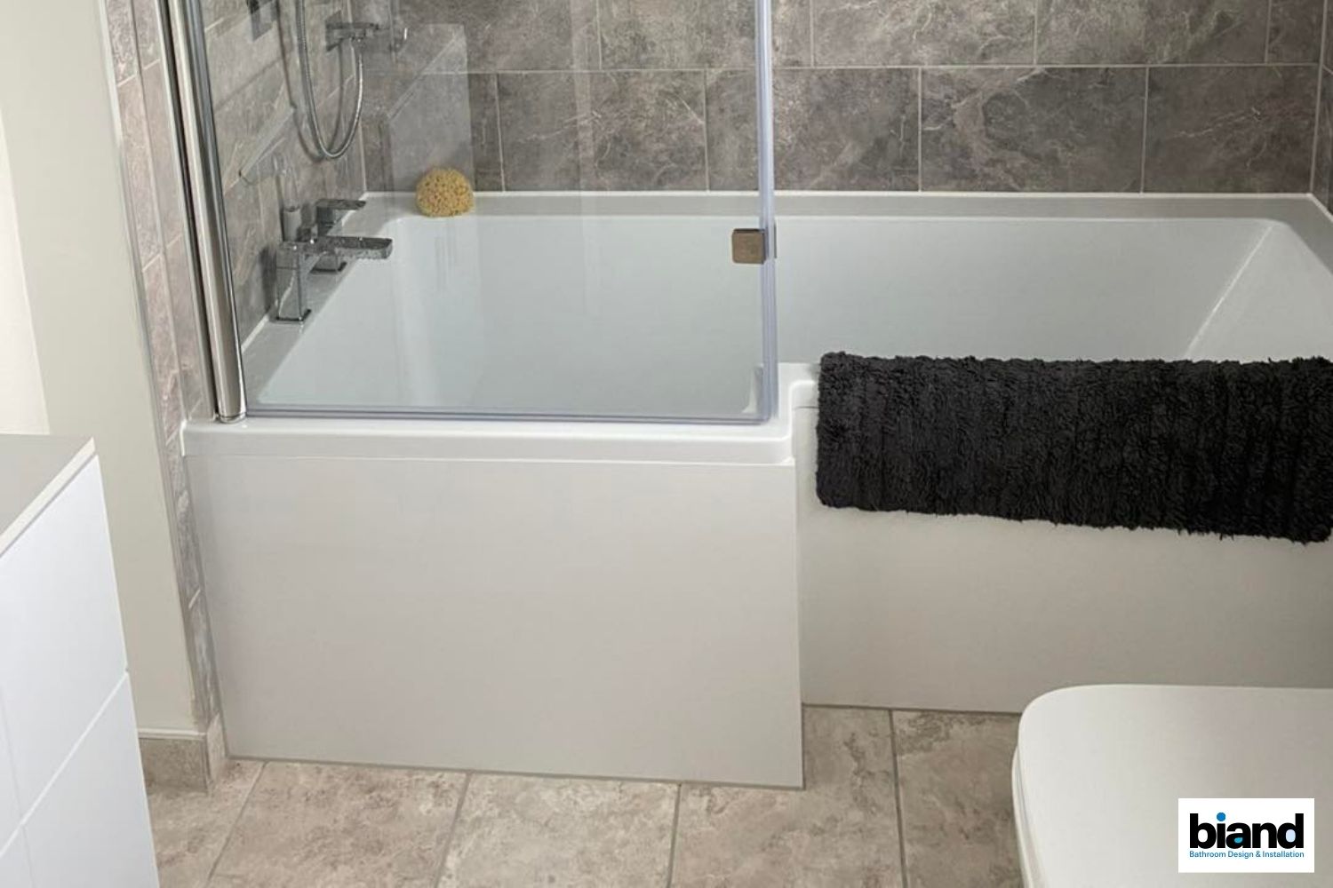 Bathtub with shower glass, grey tiled walls, and a black bath mat.
