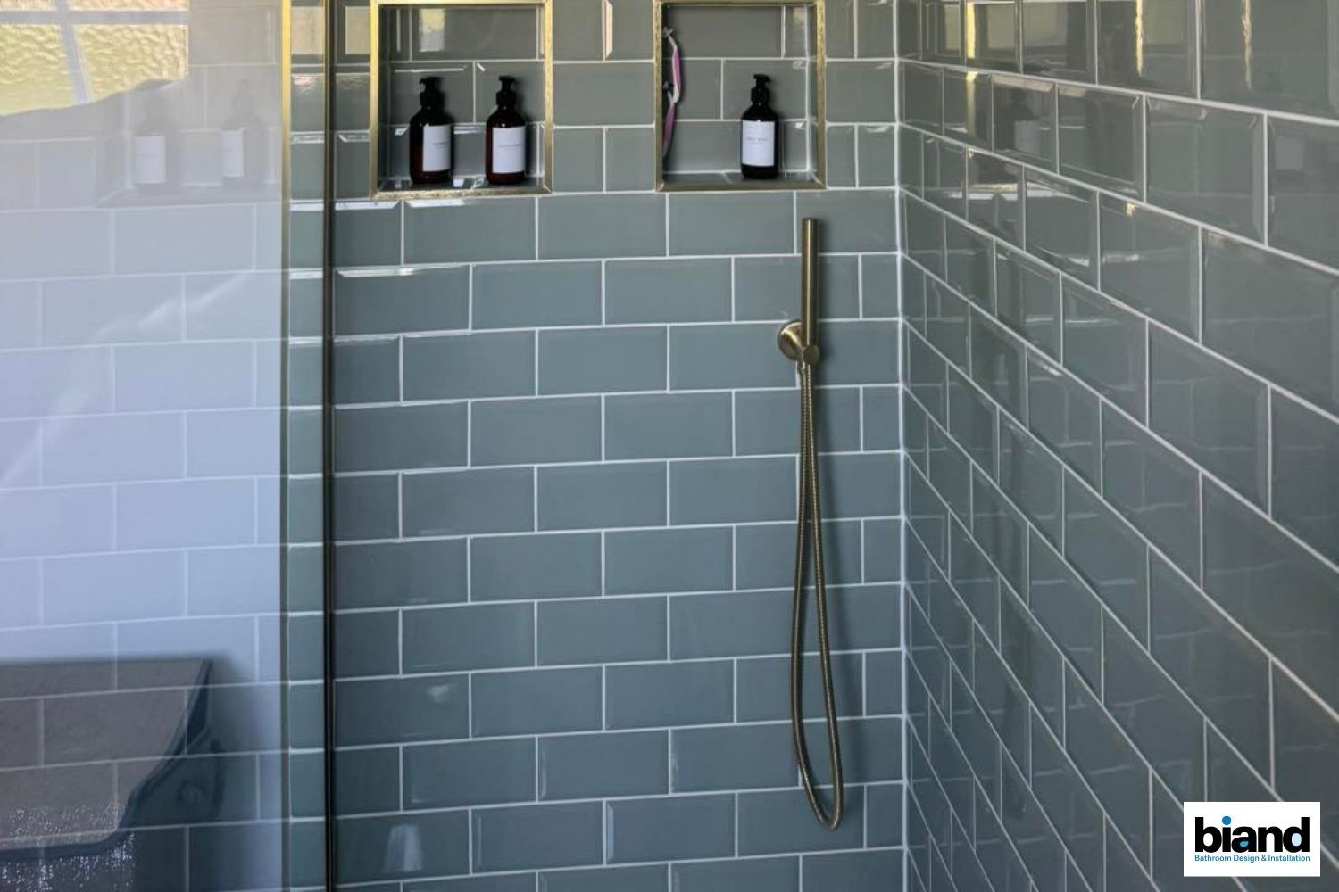 Gray tiled shower with glass door, built-in shelving for toiletries, gold fixtures.