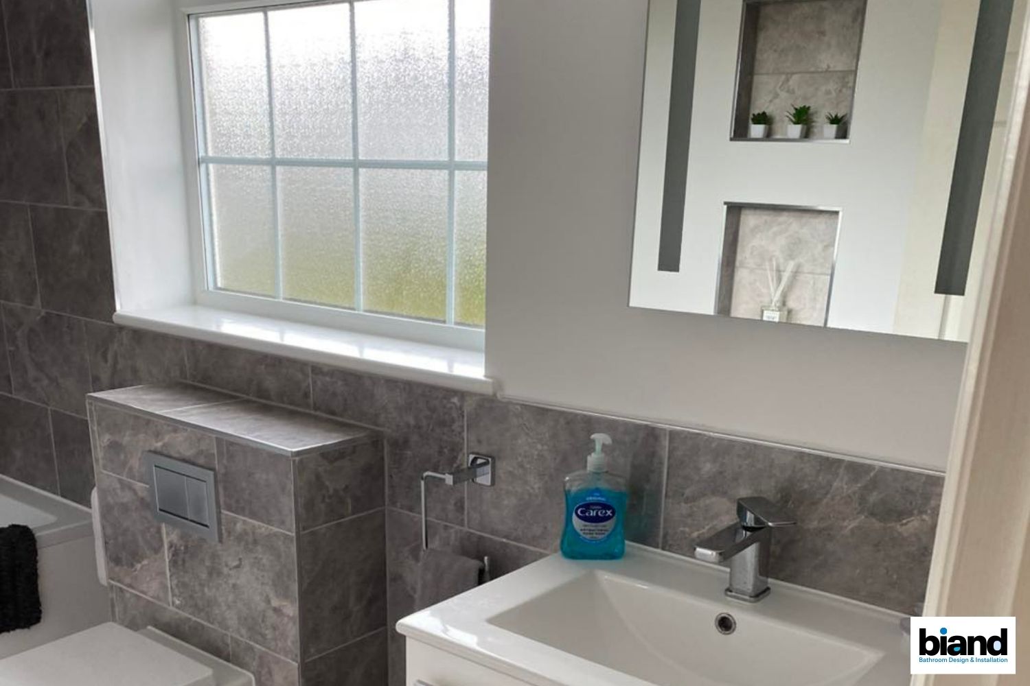 Modern bathroom with gray marble tiles, white sink, large mirror, and window.