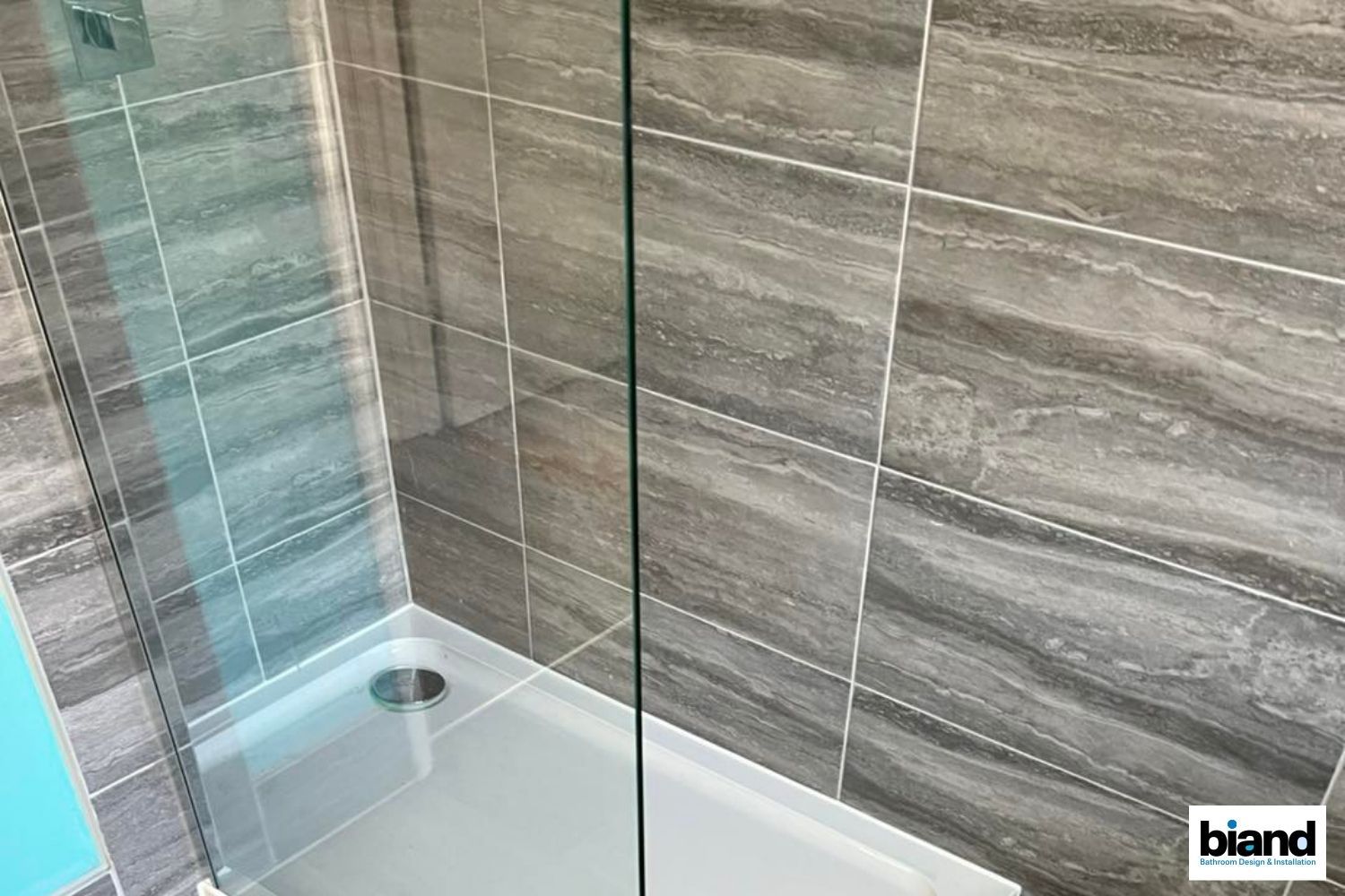 Shower stall with gray marble-look tiles, glass door, white base, and light blue wall.