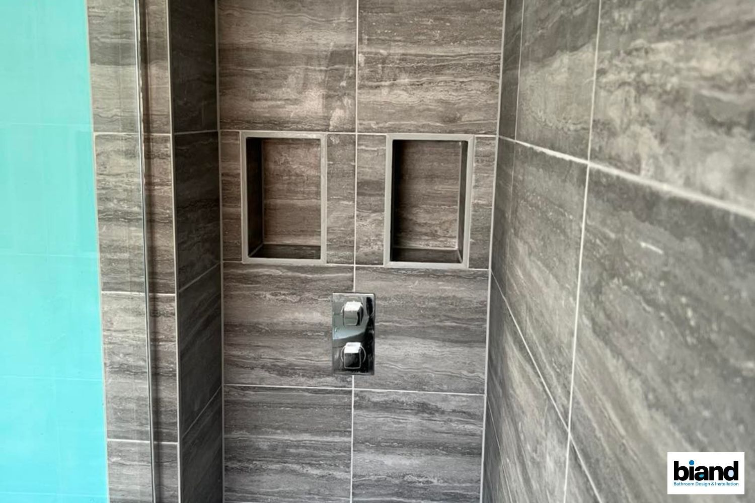 Gray tiled shower with two built-in shelves and a chrome faucet against a light blue wall.