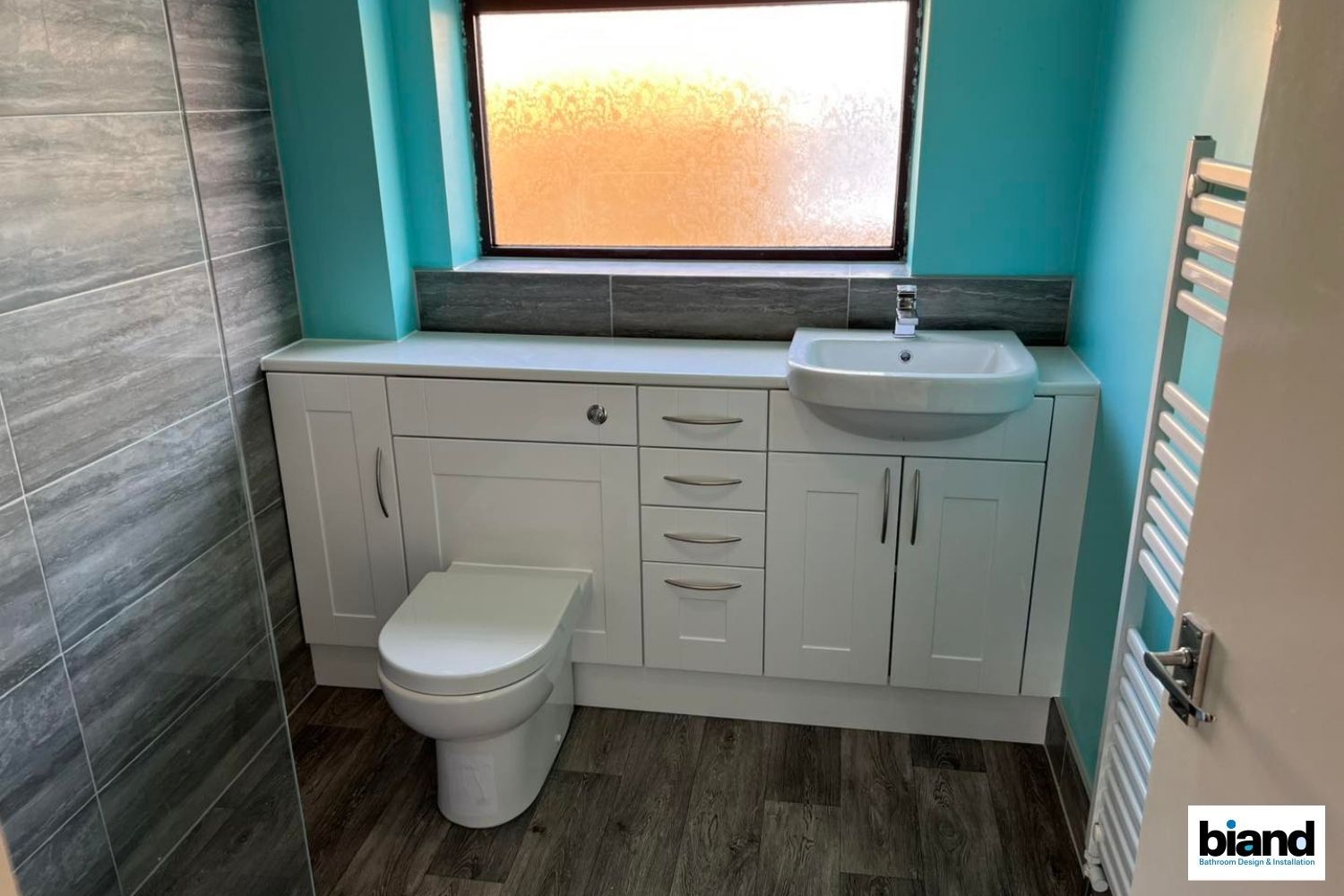 White bathroom with toilet, sink, and storage cabinets, teal wall, and gray tile accents.