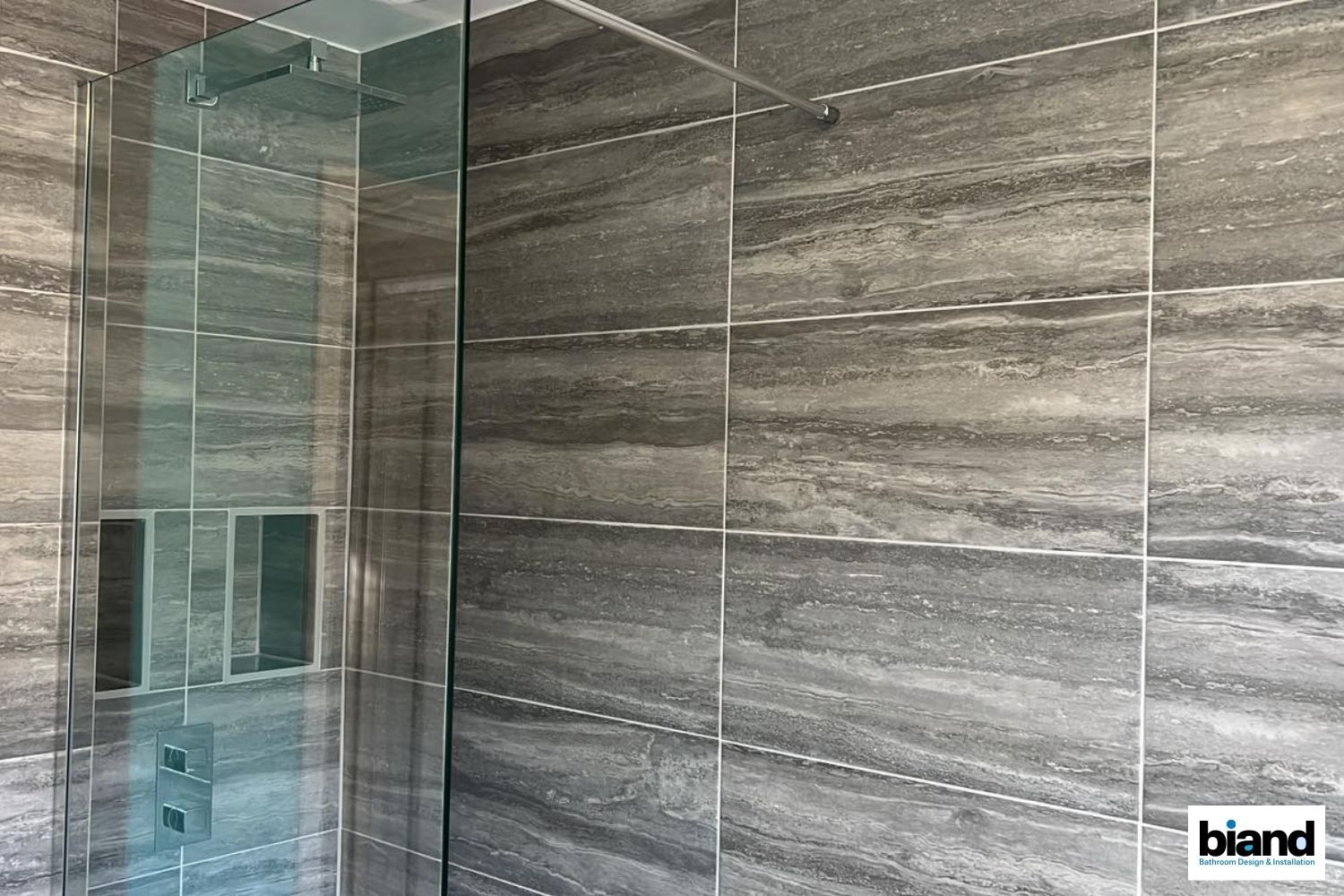 Bathroom with gray tile walls, glass shower enclosure, and built-in soap shelves.