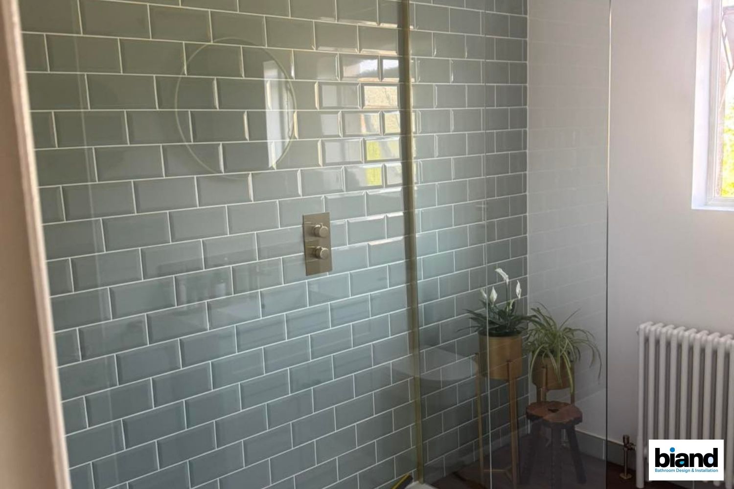 Shower with gray-green brick-style tiles, glass door, and silver fixtures; next to a plant and a stool near a window.