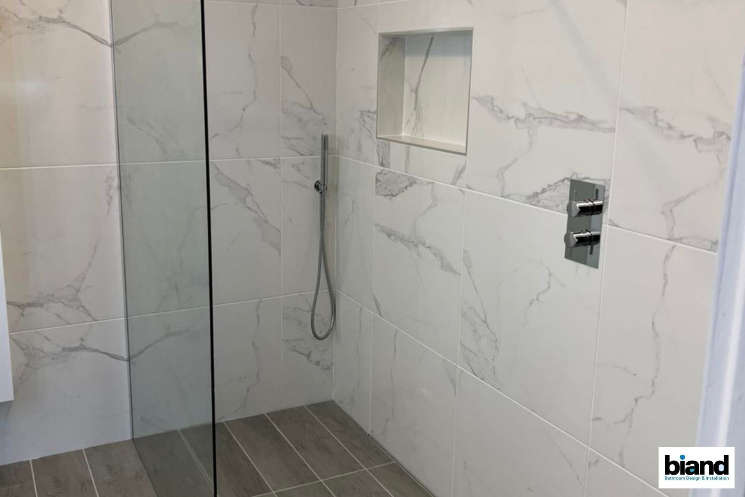 Shower with white marble-look tiles, glass door, recessed shelf, and a handheld showerhead; the floor has wood-look tiles.