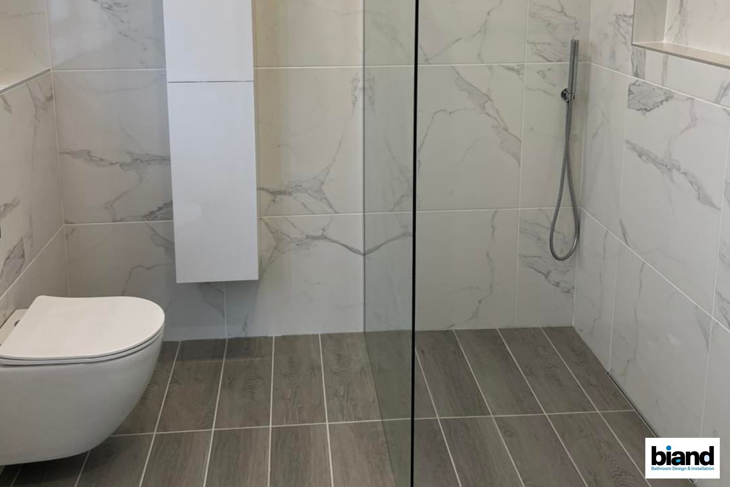 Bathroom with gray and white marble tile, shower, toilet, and white cabinet.