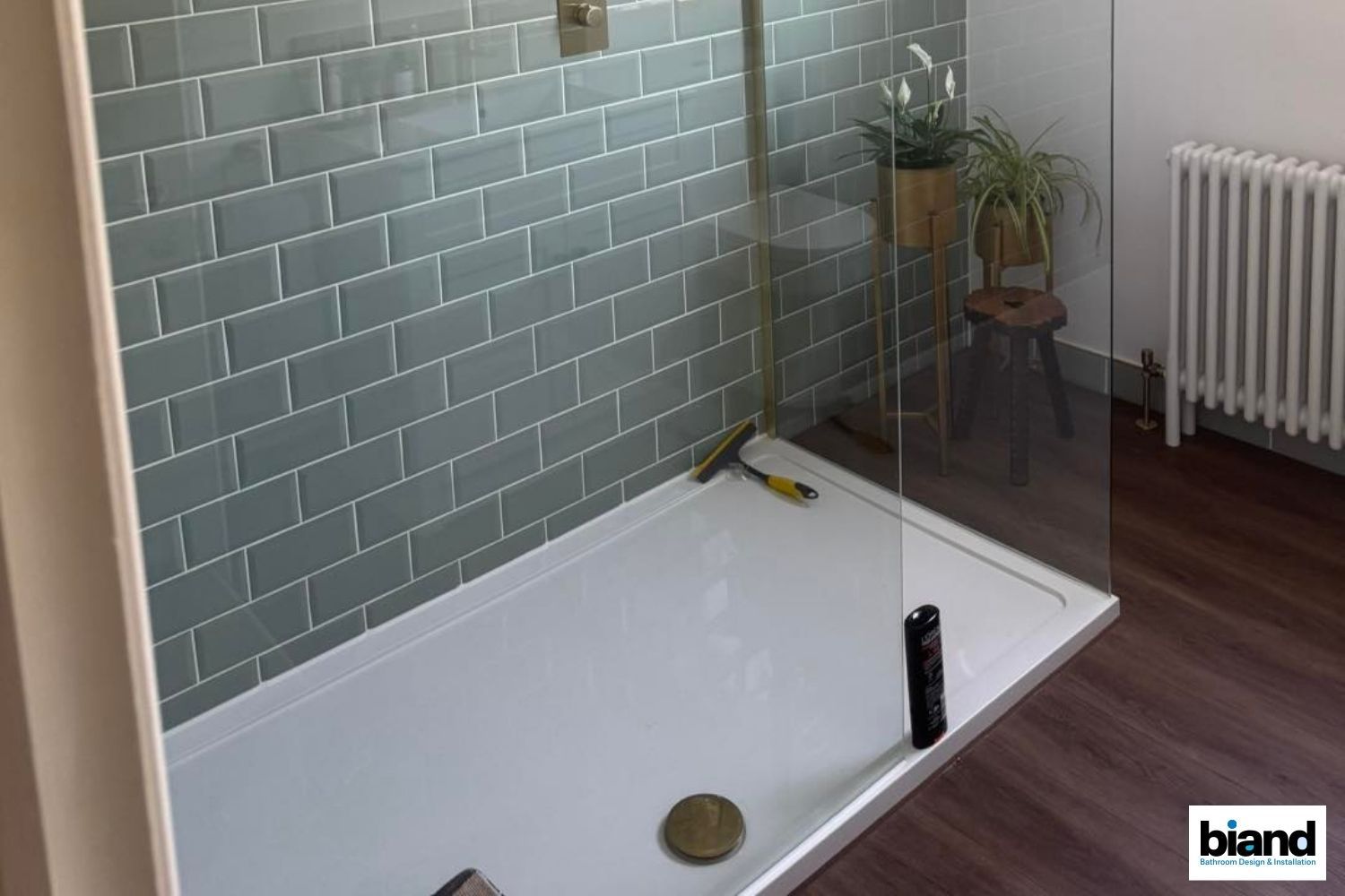 Shower with green tiled walls, a glass door, and white tray. Includes a stool and radiator.