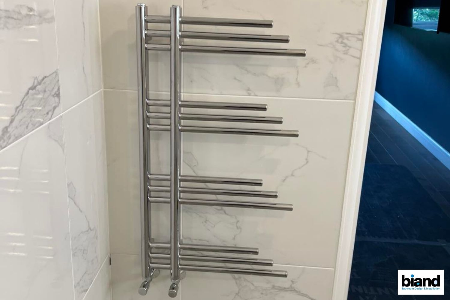 Stainless steel towel warmer mounted on white marble-look bathroom tile.
