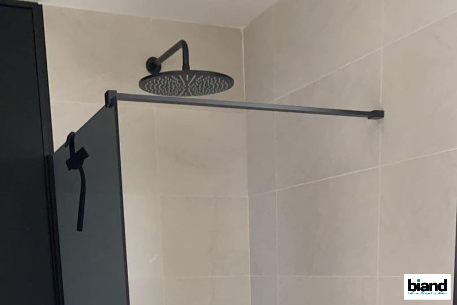 Black showerhead and rod against beige tiled wall in bathroom.