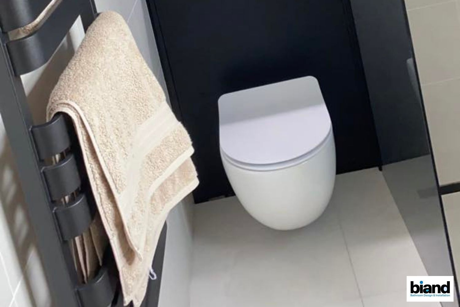White toilet next to a black towel warmer with a beige towel, in a bathroom with white tile.