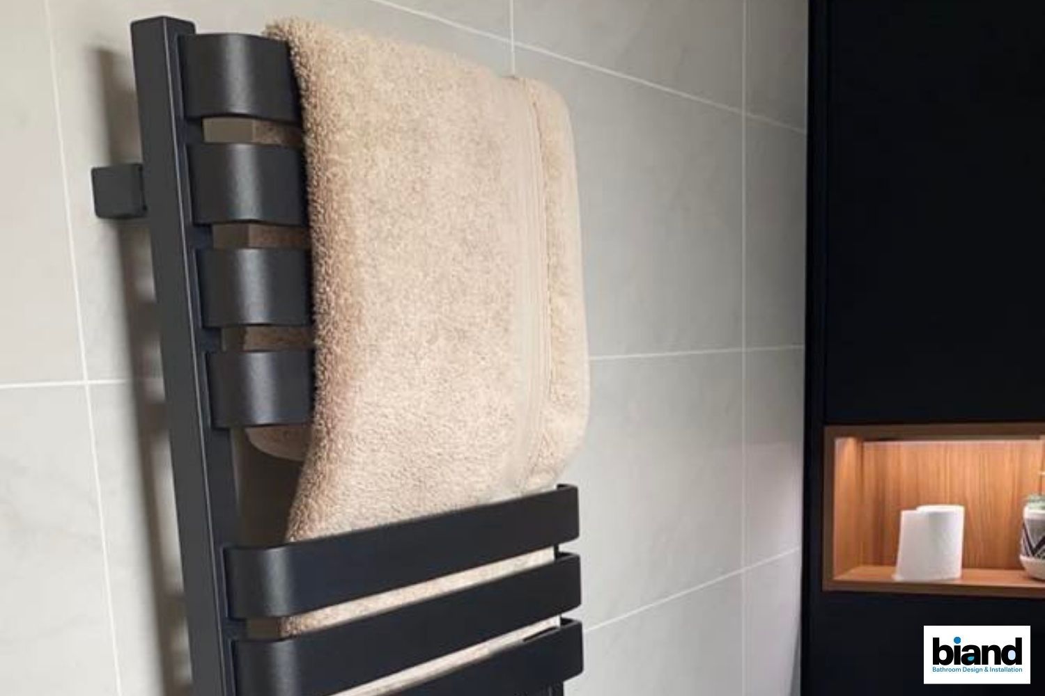 Black towel warmer with tan towel hanging on a tiled bathroom wall, wooden recessed shelf on the right.
