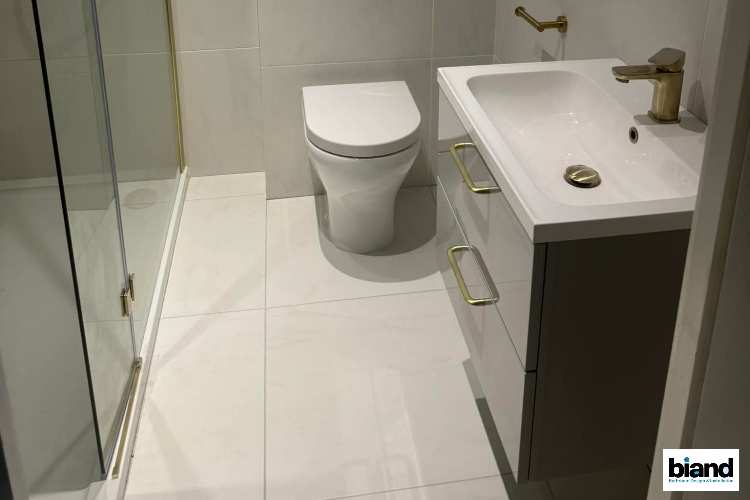 A modern bathroom with white tiles, a toilet, vanity with gold fixtures, and a shower.