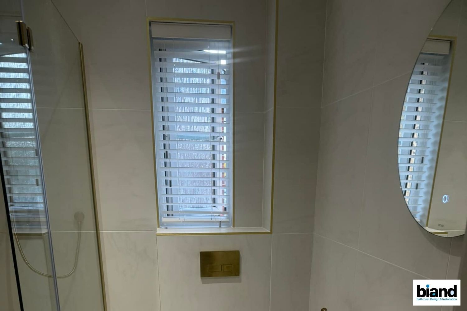 Bathroom with cream tile walls, gold accents, and a narrow window with blinds. Two mirrors reflect the scene.