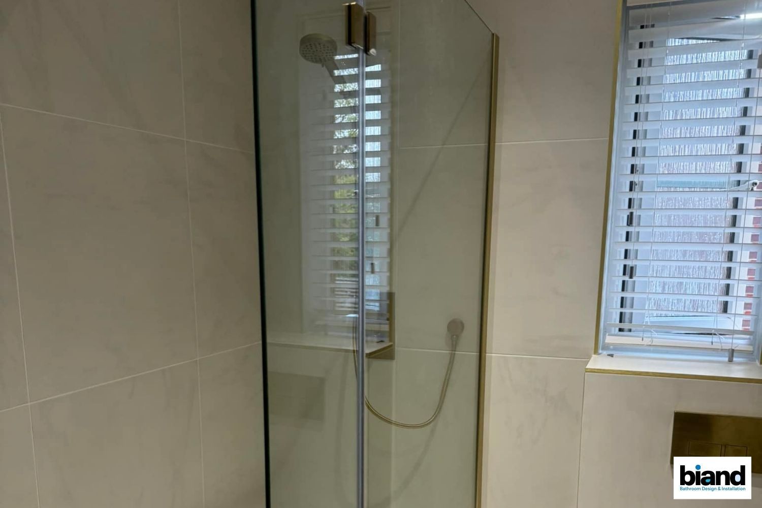 Shower stall with gold trim, beige tile, and a window with white blinds.