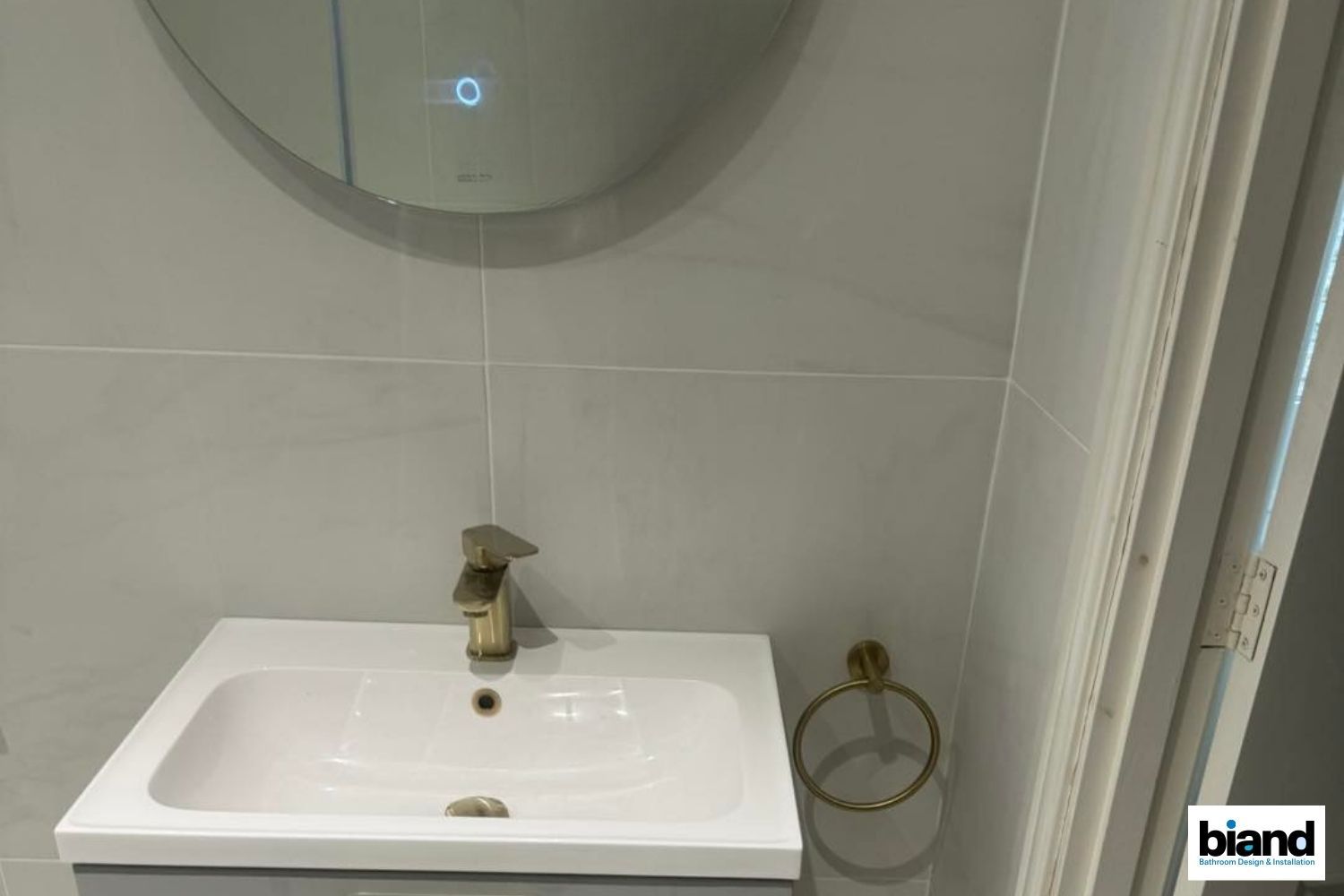 Modern bathroom with a white sink, gold faucet, and round mirror. Gray tiles and a gold towel ring.