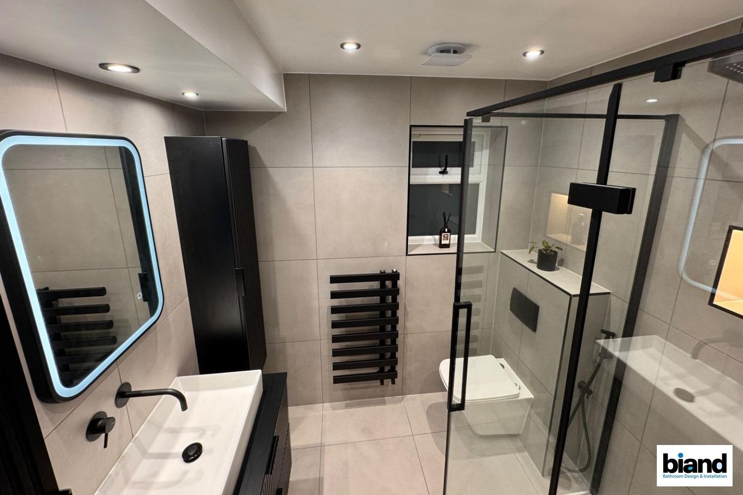 Modern bathroom with black fixtures, gray tile, glass shower, and illuminated mirror.