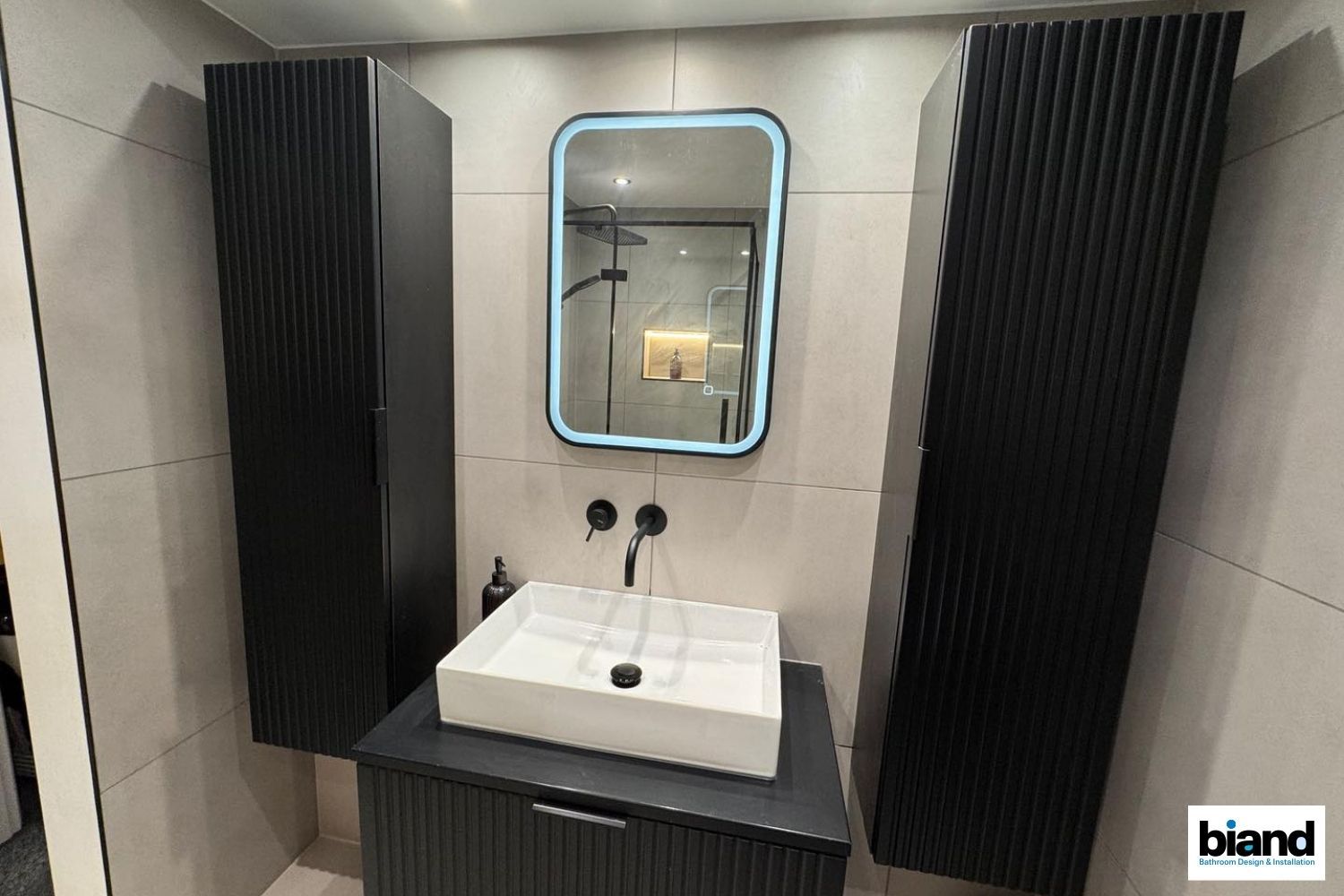 Modern black and white bathroom with tall cabinets, a square sink, and illuminated mirror.