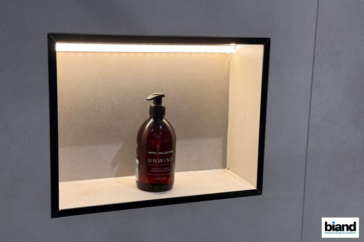 A recessed shelf with a brown soap bottle and warm lighting in a gray tiled wall.
