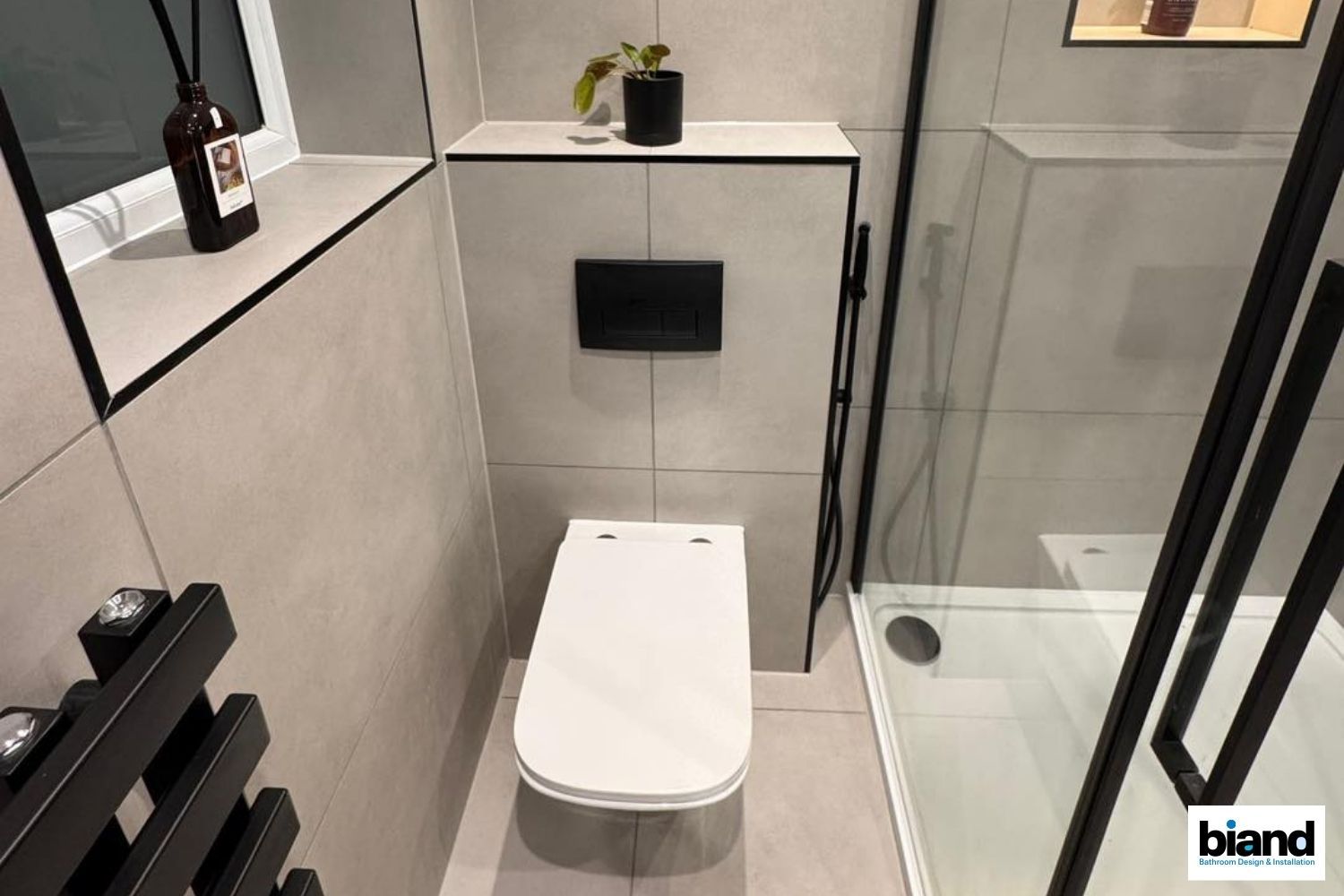 Modern bathroom: toilet, built-in cabinet, black fixtures, shower, small plant on top, and grey tile.