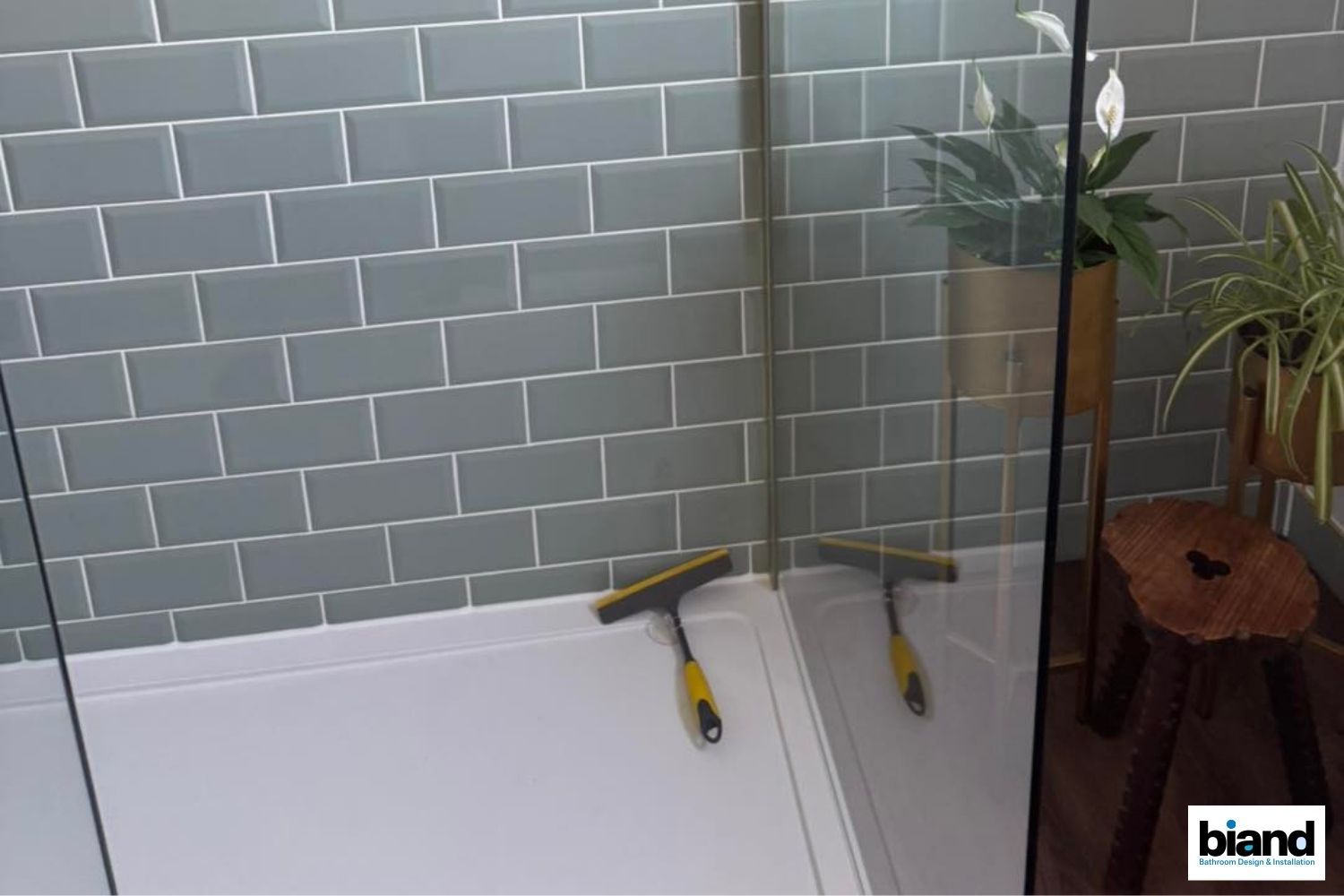 Shower with blue-gray tiled walls and glass door. A squeegee sits in the white shower base. A wooden stool and plants are in the corner.