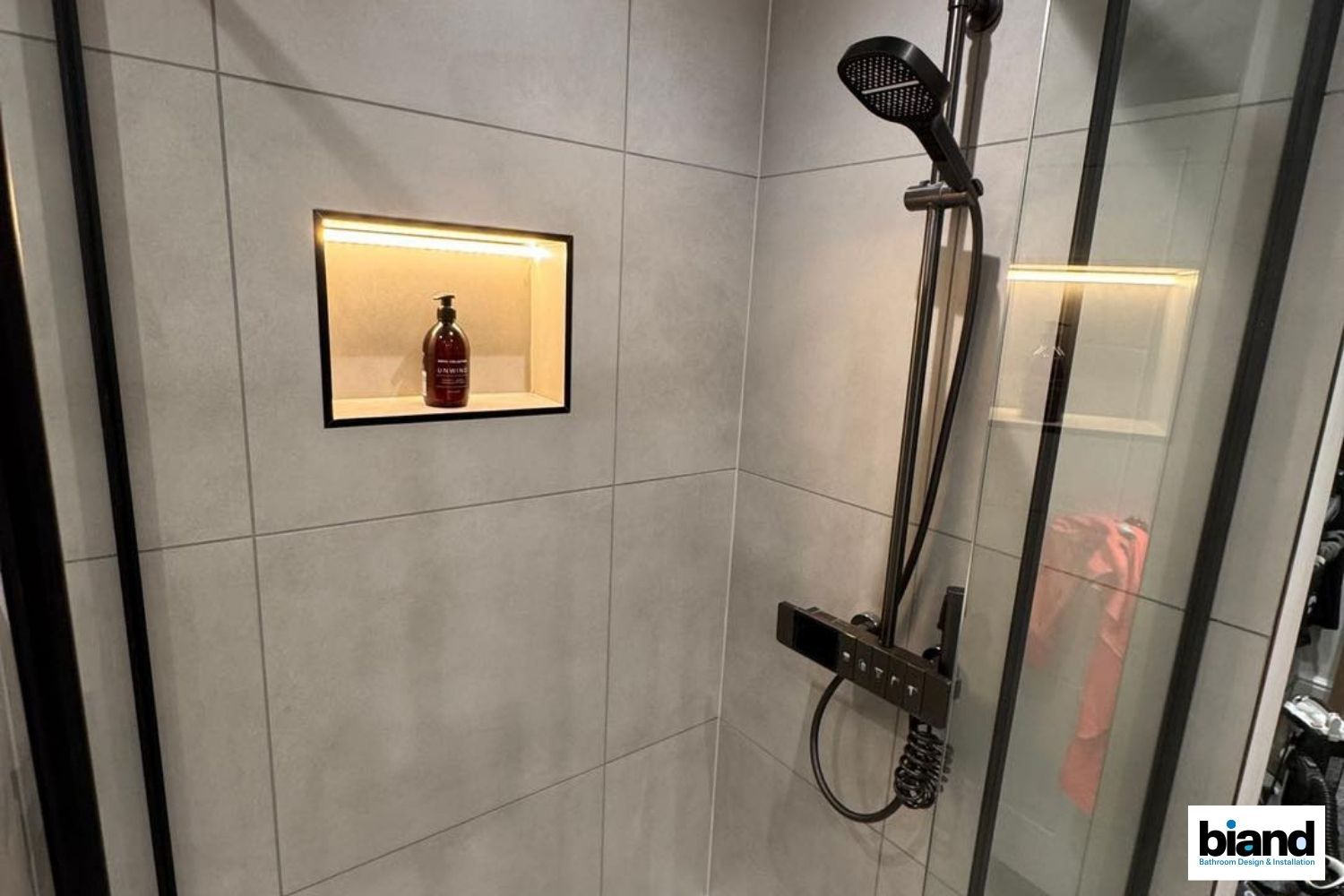 Shower with gray tiles, built-in shelf with a bottle, and black shower fixtures against a glass enclosure.