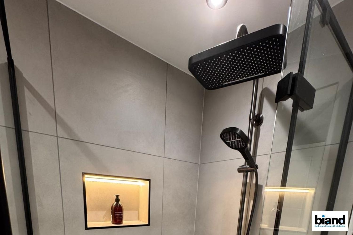 Modern shower with gray tile walls, black fixtures, and recessed shelf with illuminated bottle.