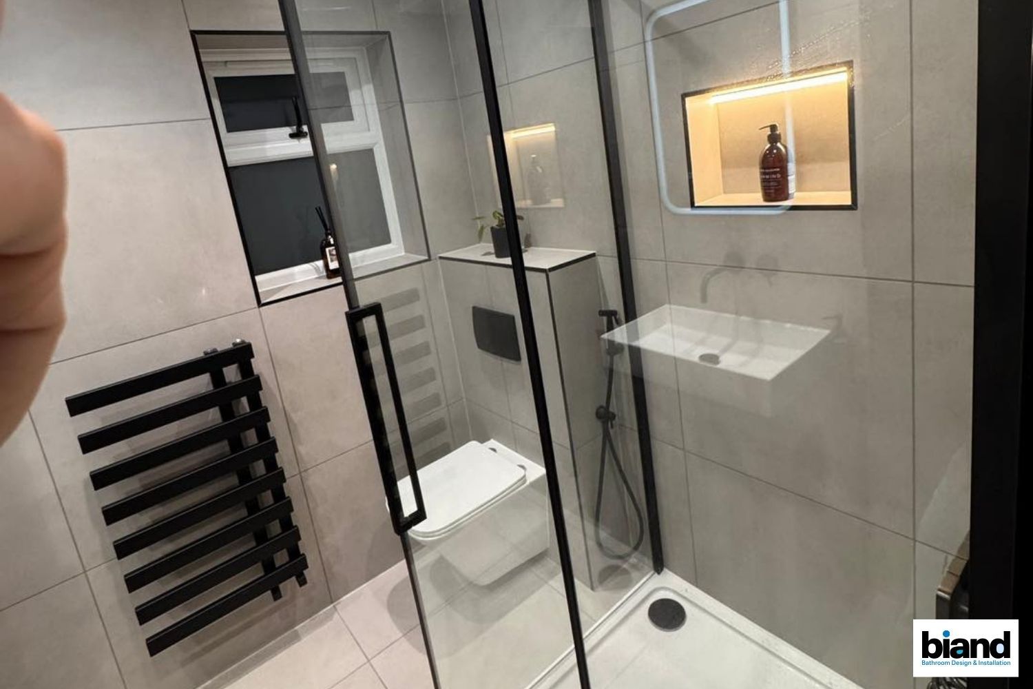Modern bathroom with a glass shower, toilet, and black fixtures. Gray tiled walls and a small window.