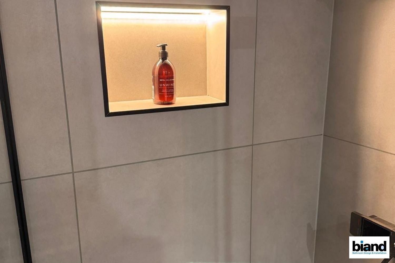 Recessed shower shelf with a bottle of soap, lit from above, on a tiled wall.