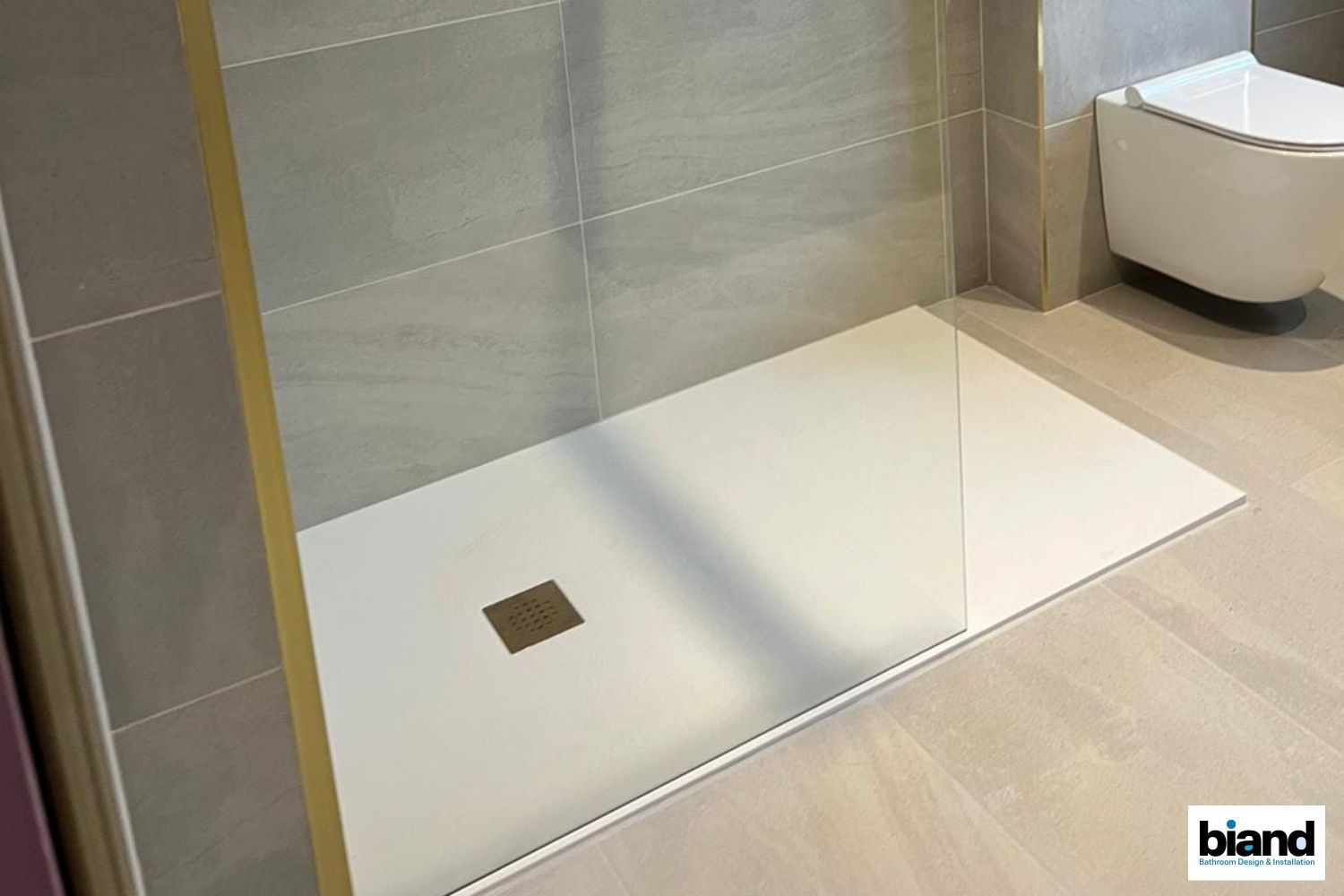 Modern white shower floor with a drain, next to a clear glass panel, and a wall-mounted toilet.