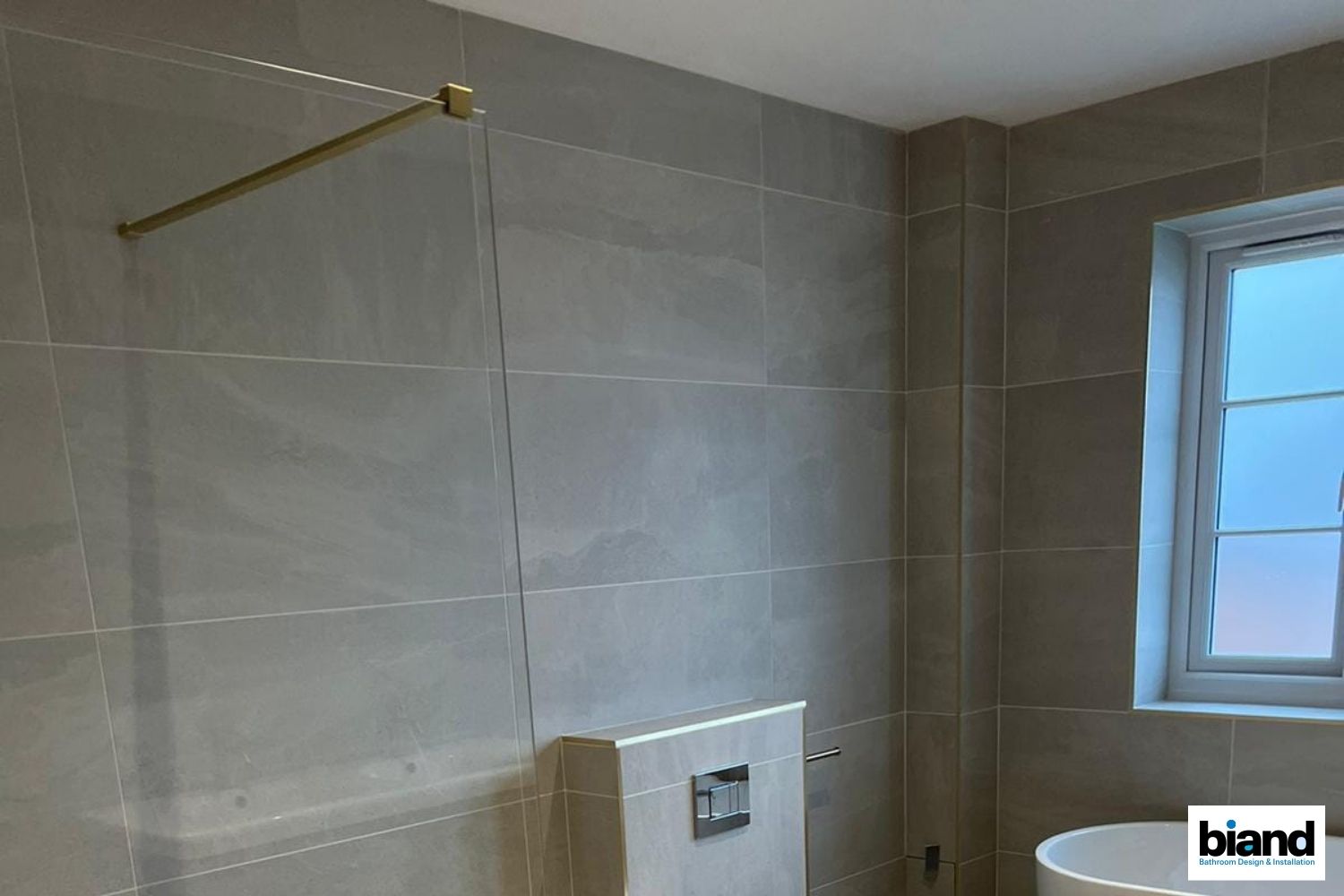 Bathroom with gray tiled walls, gold shower rod, and a window.