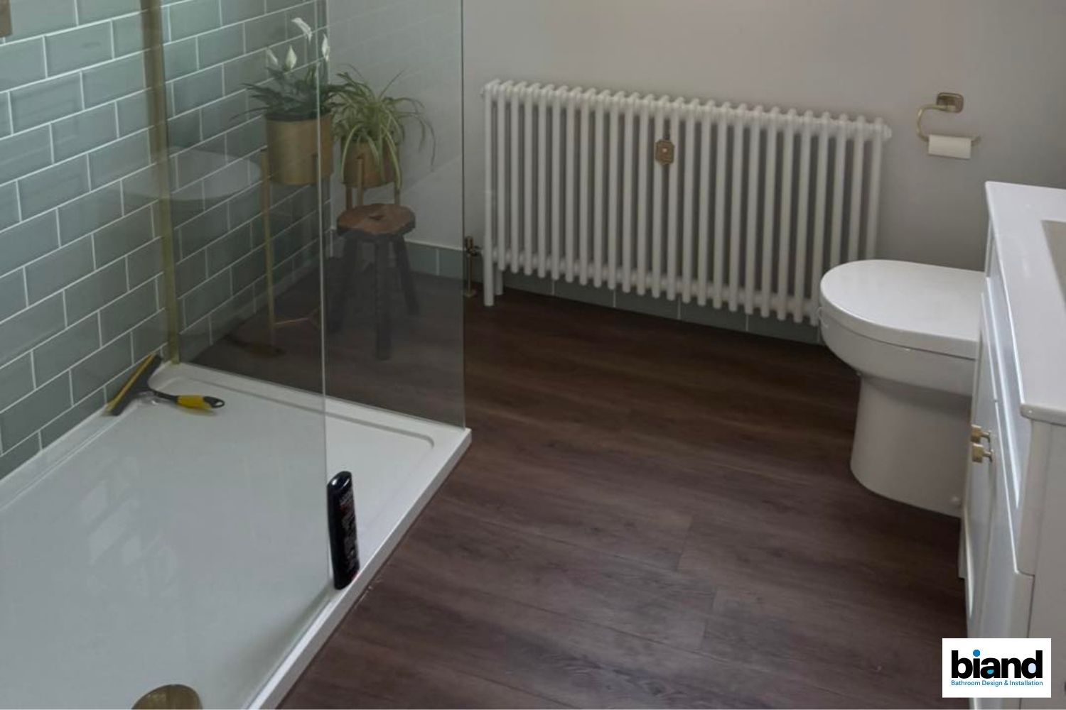 A modern bathroom with teal tiled walls, a white shower, radiator, toilet, and dark wood-look flooring.