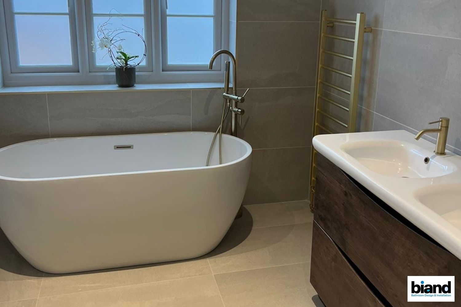 Modern bathroom with a white tub, vanity, gold fixtures, and a window.