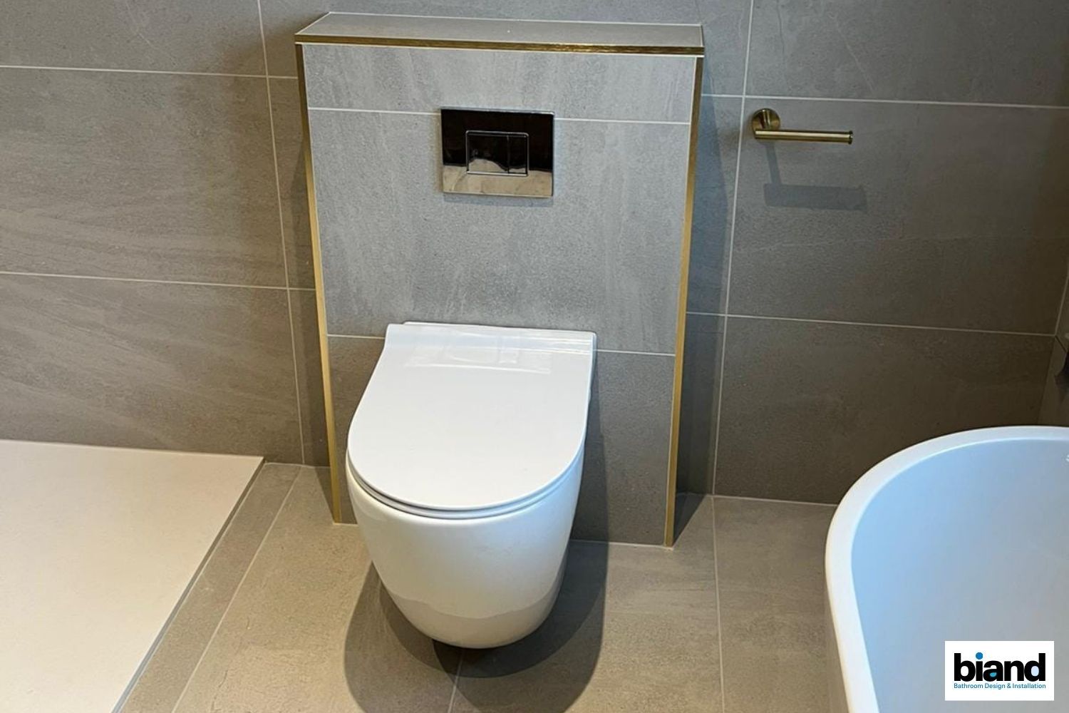 Modern bathroom with a wall-mounted toilet, gray tiles, gold trim, and fixtures.