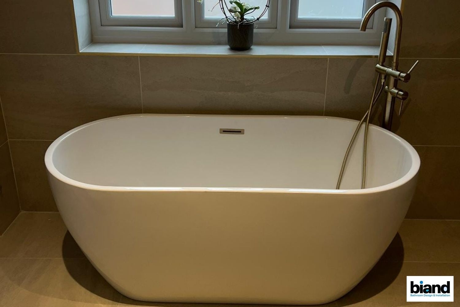 Oval white bathtub in a neutral-toned bathroom with window in the background, silver faucet.
