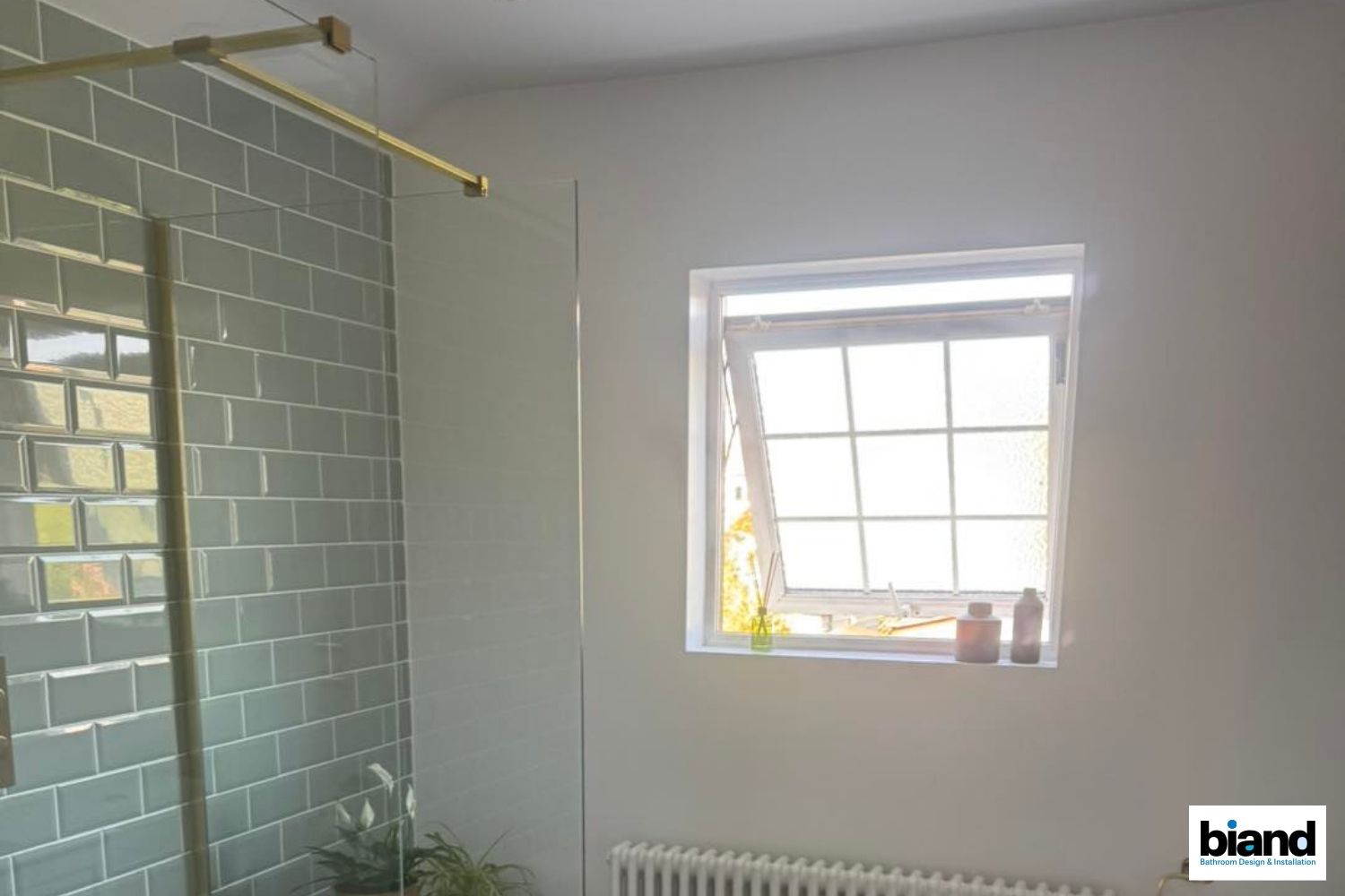 Bathroom with green tiled wall, shower glass, open window with sunlight, and neutral-colored walls.