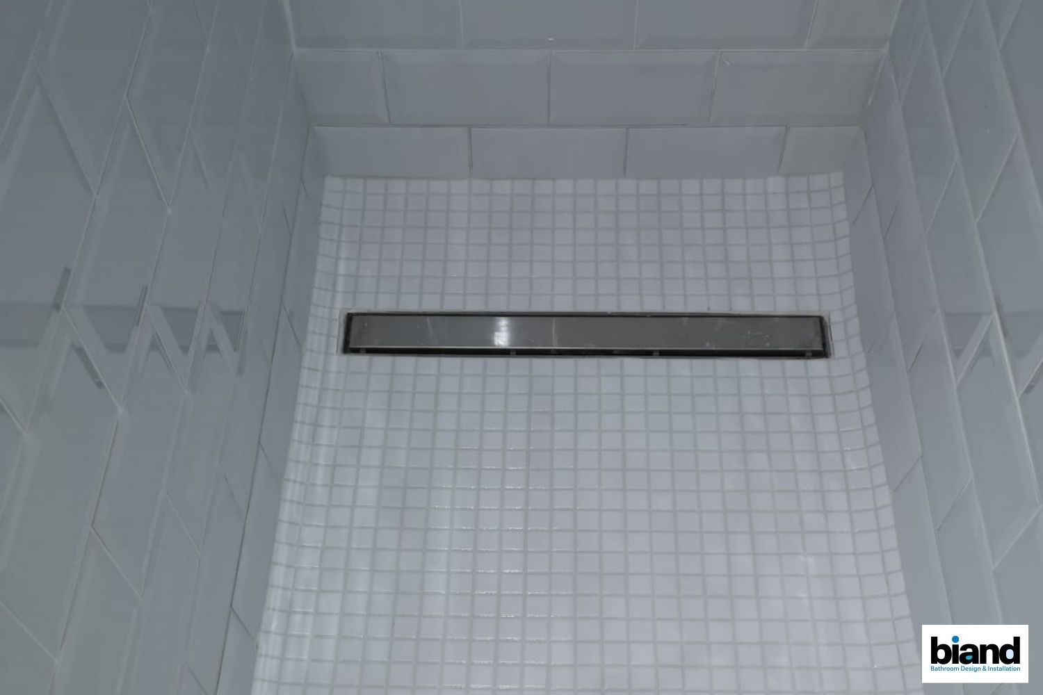 A shower interior with white tiled walls and floor, featuring a long, rectangular drain.