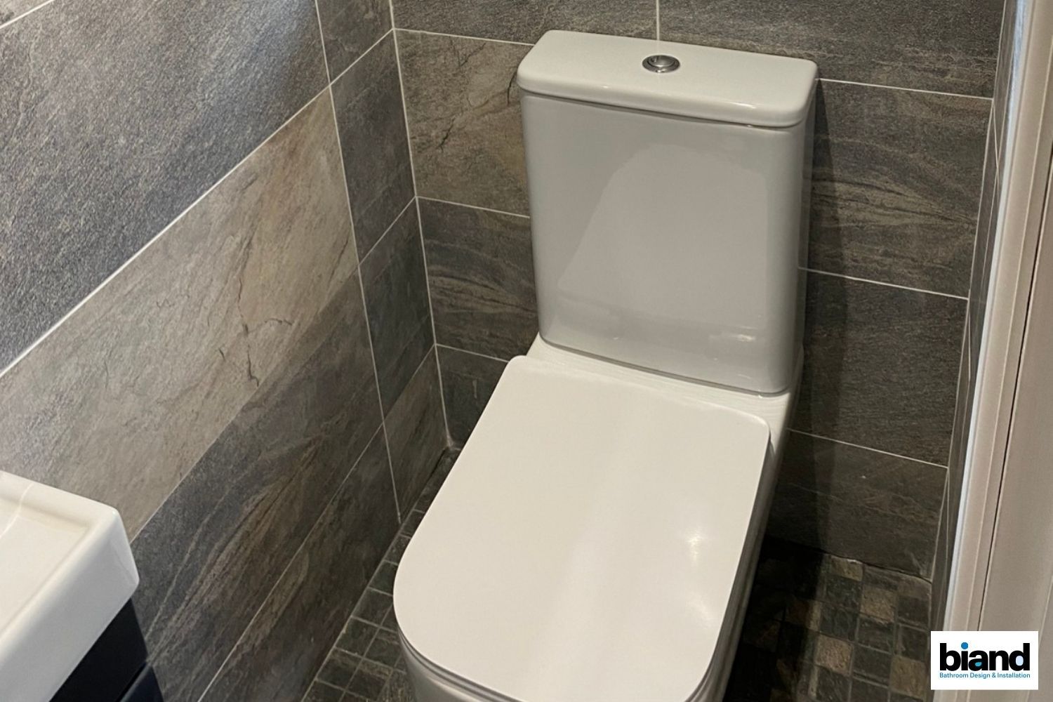 Toilet in a bathroom with gray and brown tiled walls. White toilet seat and tank, neutral colors.